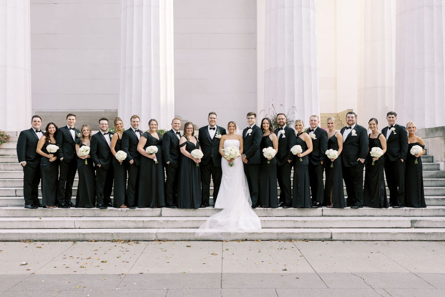 Large wedding party stands in line along white columns of a grand building during formal photo session.