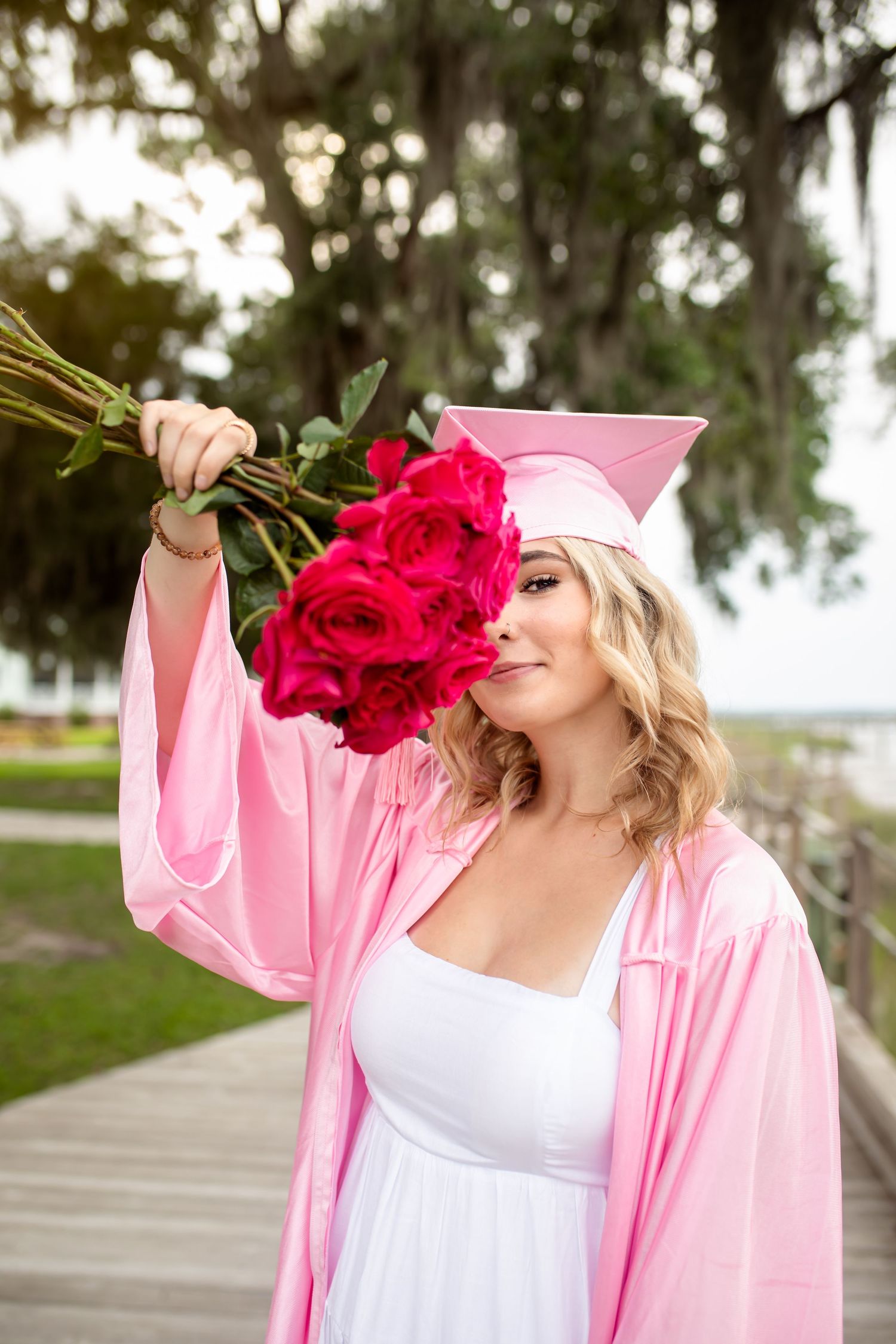 A graduate in pink cap and gown smells a bouquet of red roses while standing outdoors on a boardwalk.