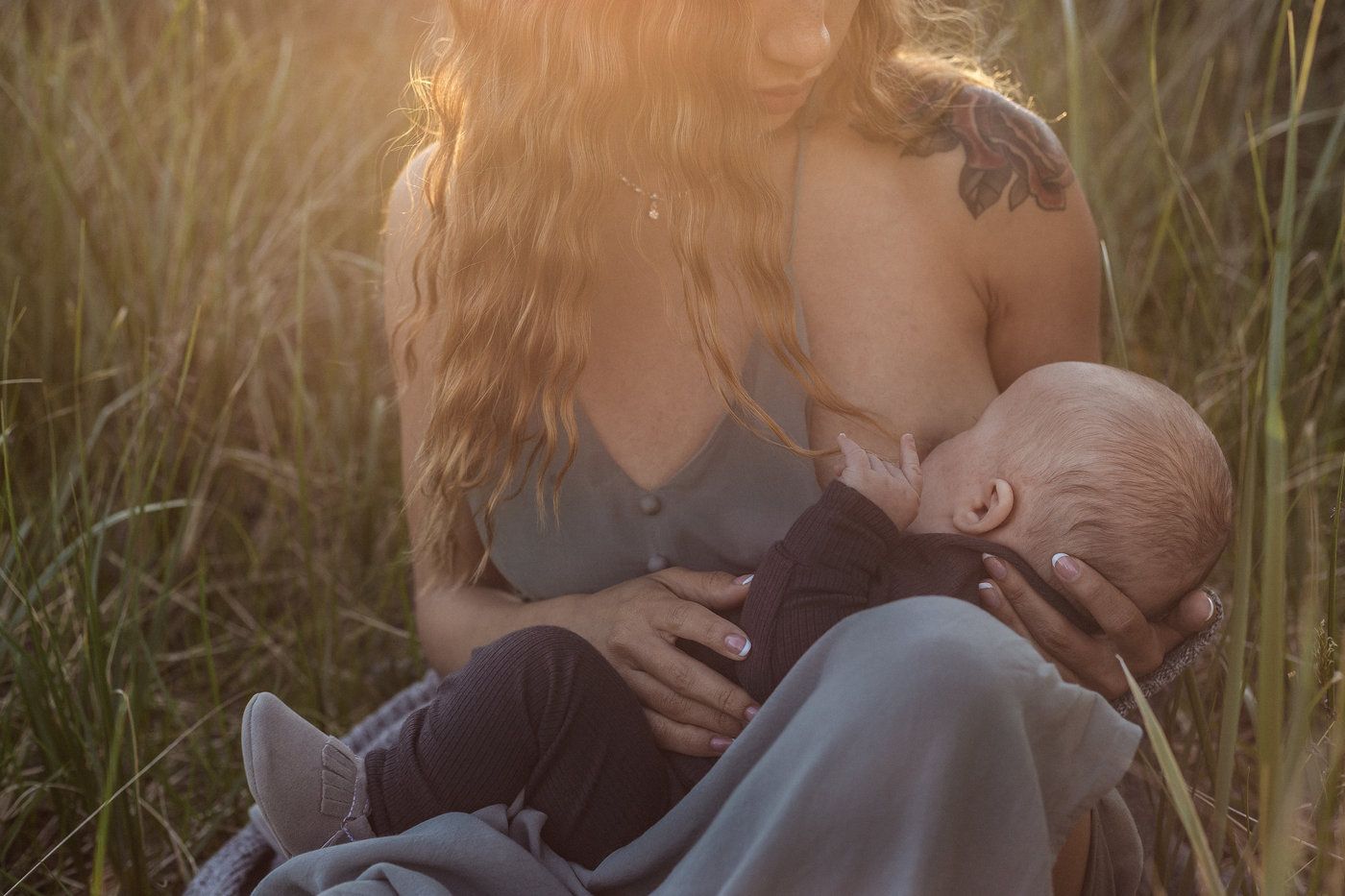 Mother breastfeeding baby in tall grass at sunset with golden light filtering through the natural setting.