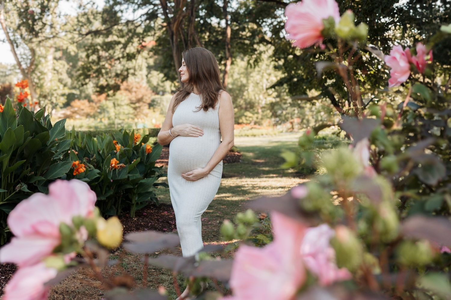 An expectant mother in white stands among blooming pink roses in a garden setting.