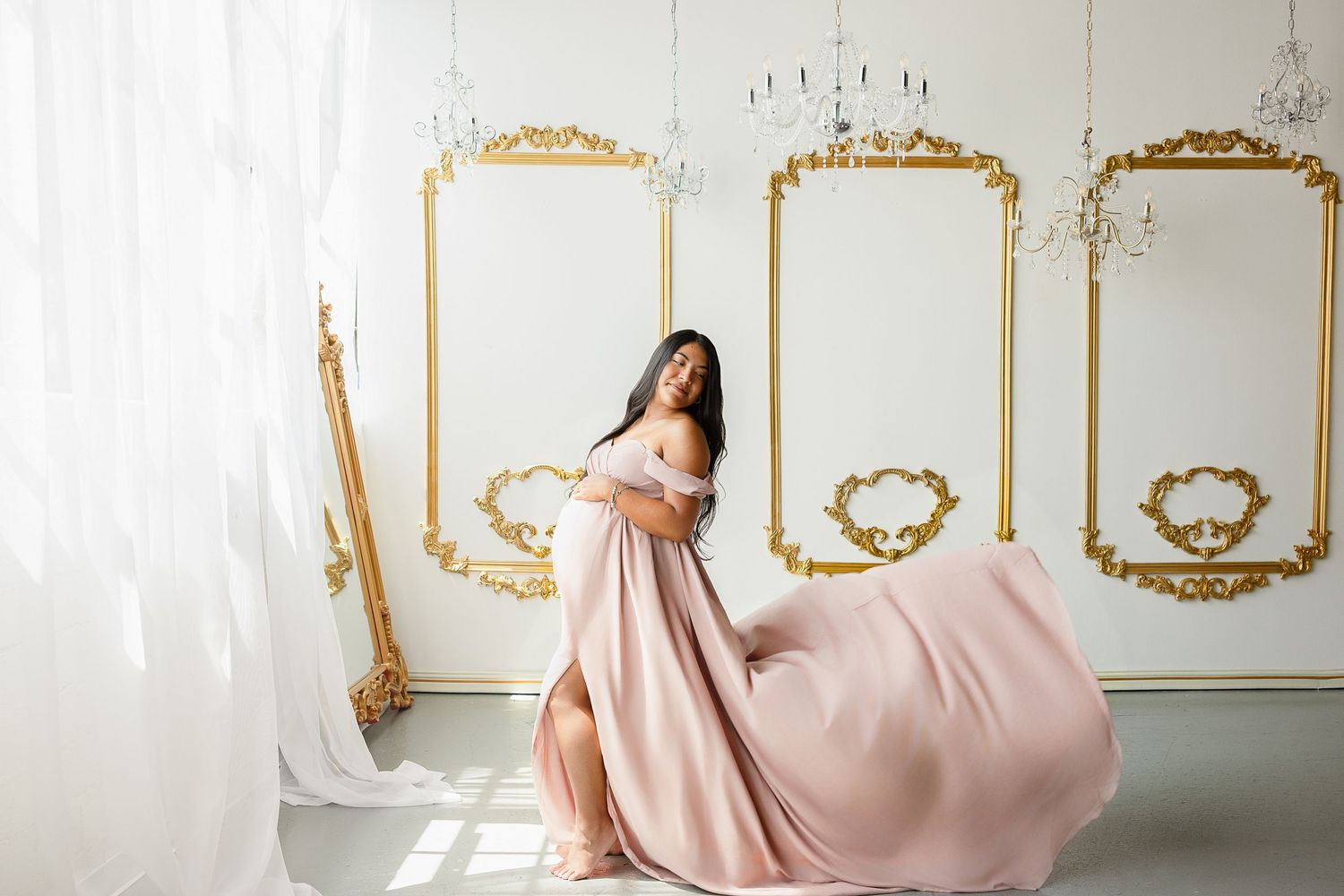 Woman in flowing pink off-shoulder gown poses gracefully in elegant white and gold ornate room.