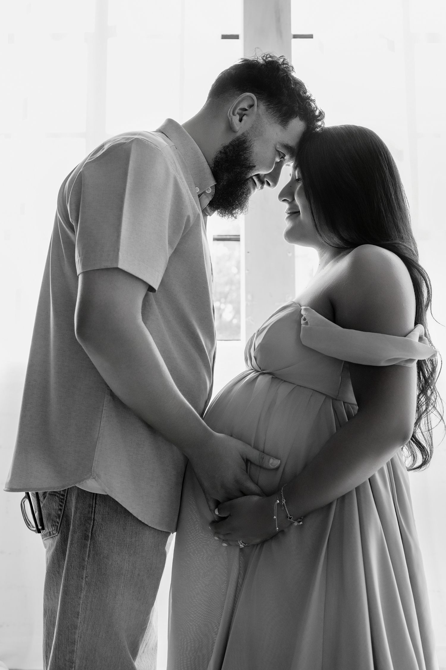 Black and white photo of a couple holding hands, the woman visibly pregnant, touching foreheads in a tender maternity moment.