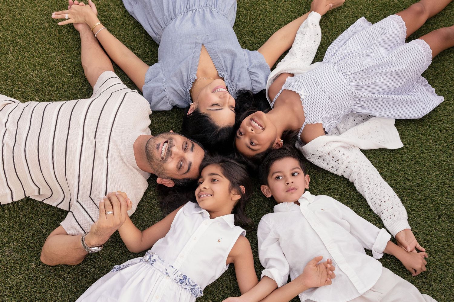 Family of five lying on green grass in a circle, smiling up at camera, wearing white and blue outfits.
