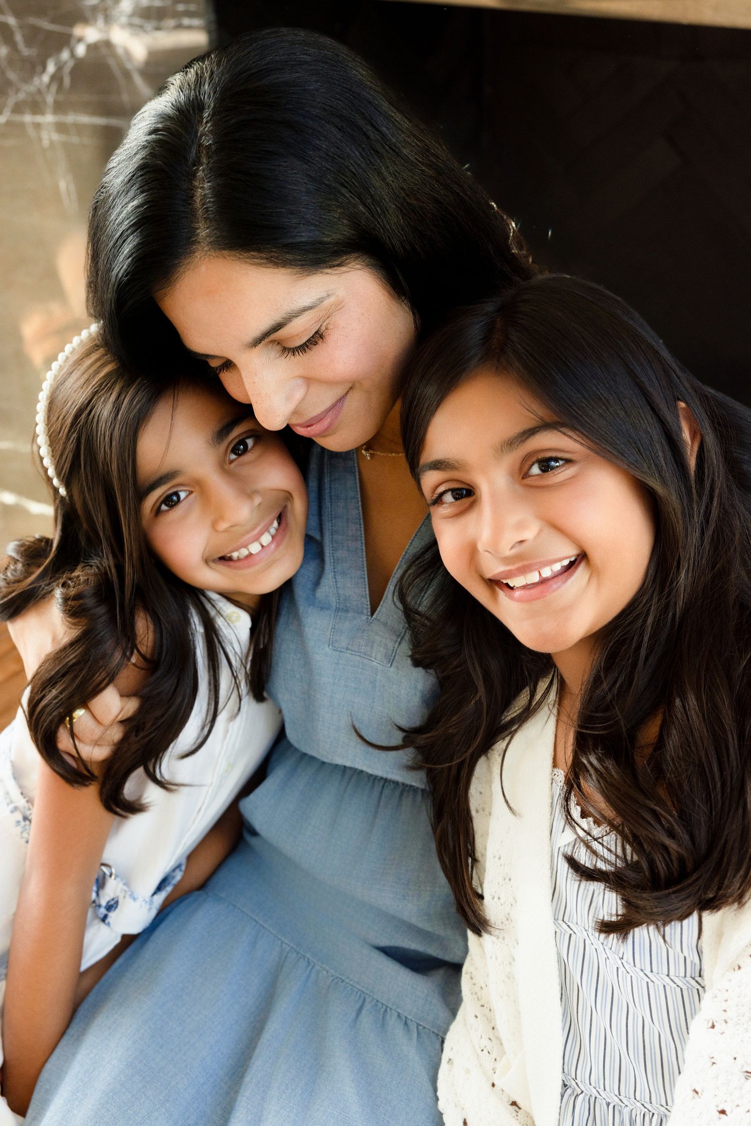 A woman smiles warmly with two young girls, all with dark hair, posing together in a cheerful family portrait.