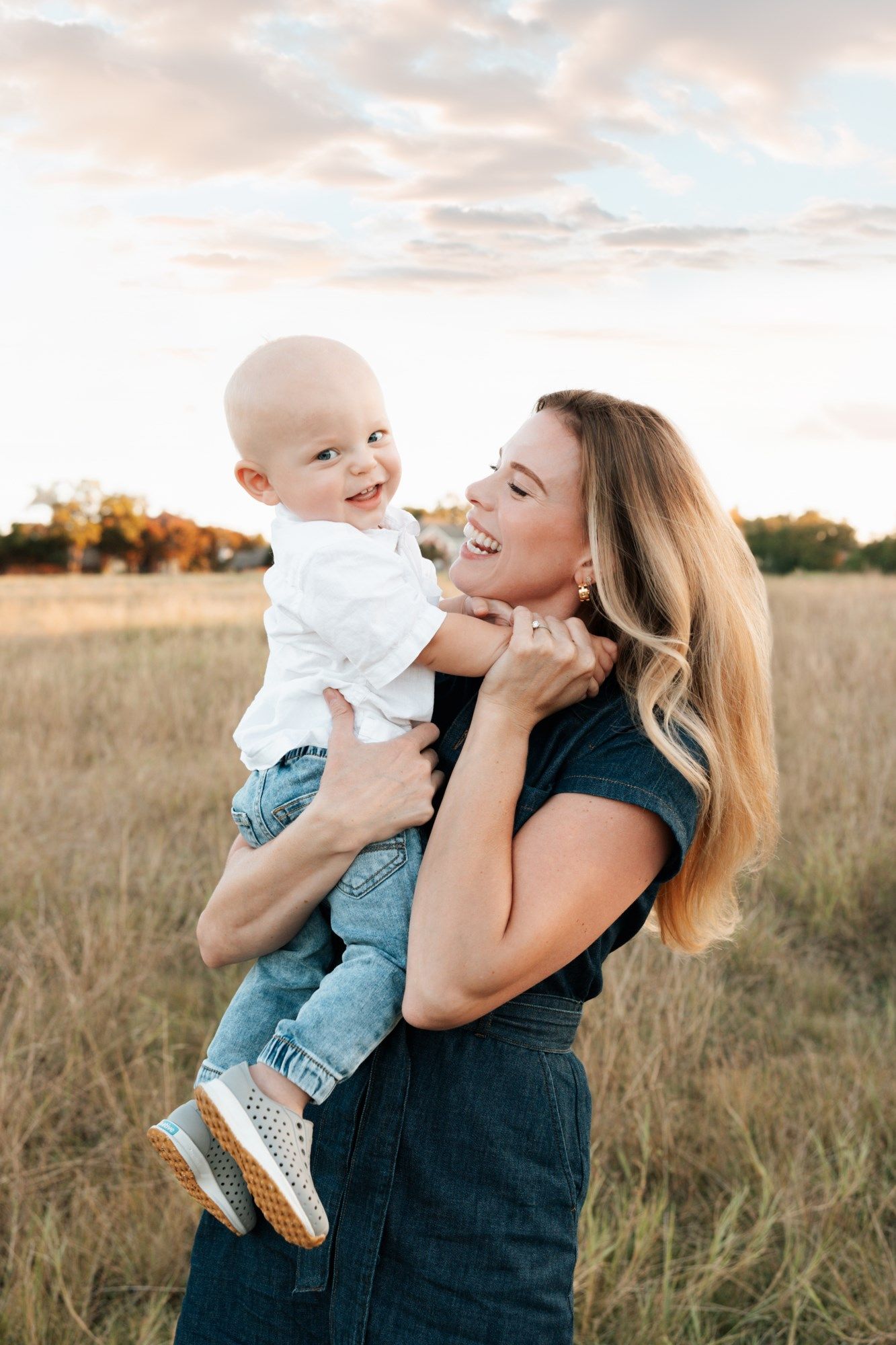 A smiling mother holds her happy baby boy in a sunlit field, both laughing joyfully during a golden hour outdoor photo session.