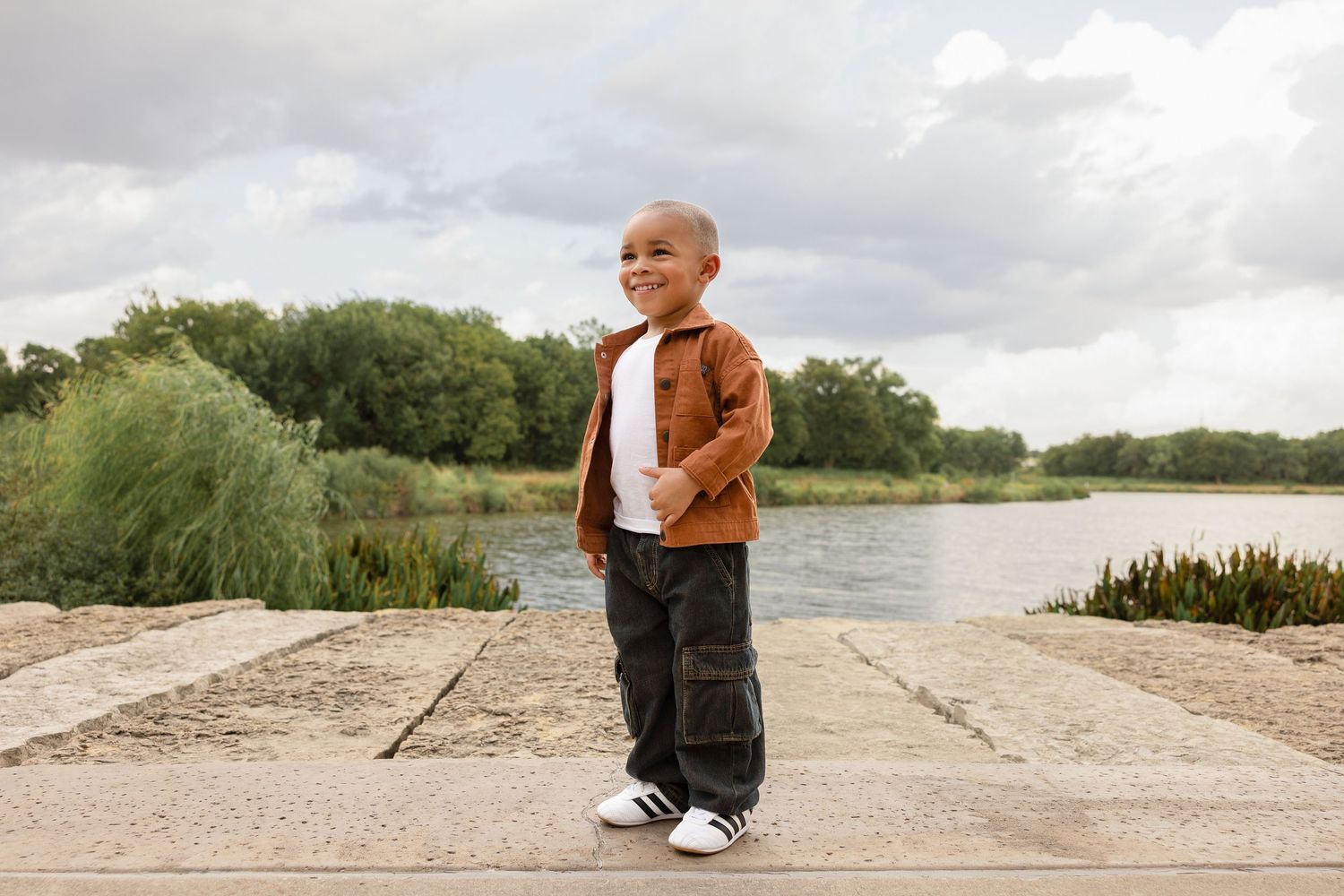A young boy in a brown jacket and black pants stands confidently outdoors near a lake with lush green trees behind him.