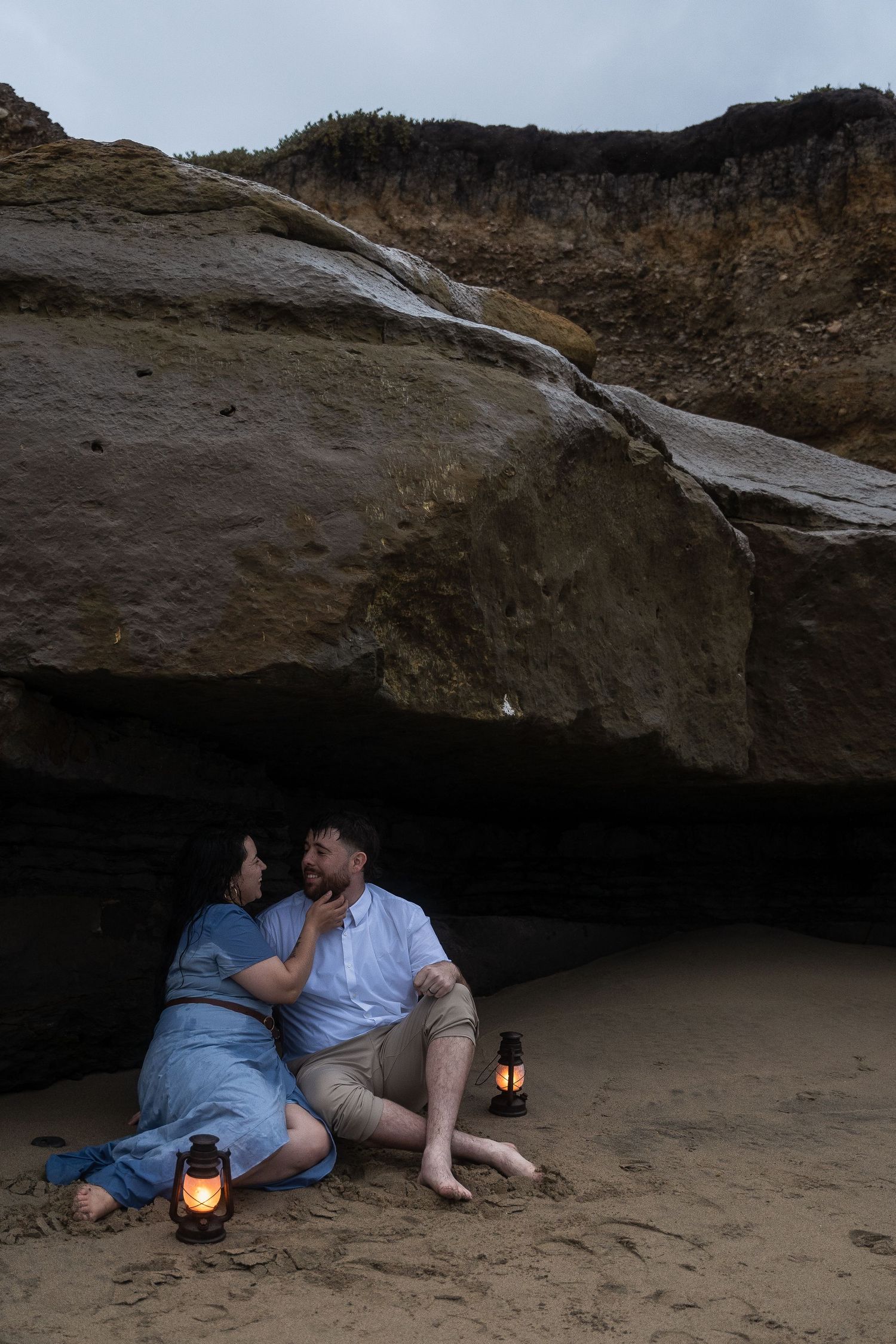 Couple sharing romantic moment in coastal cave at dusk with glowing lanterns on sandy beach.
