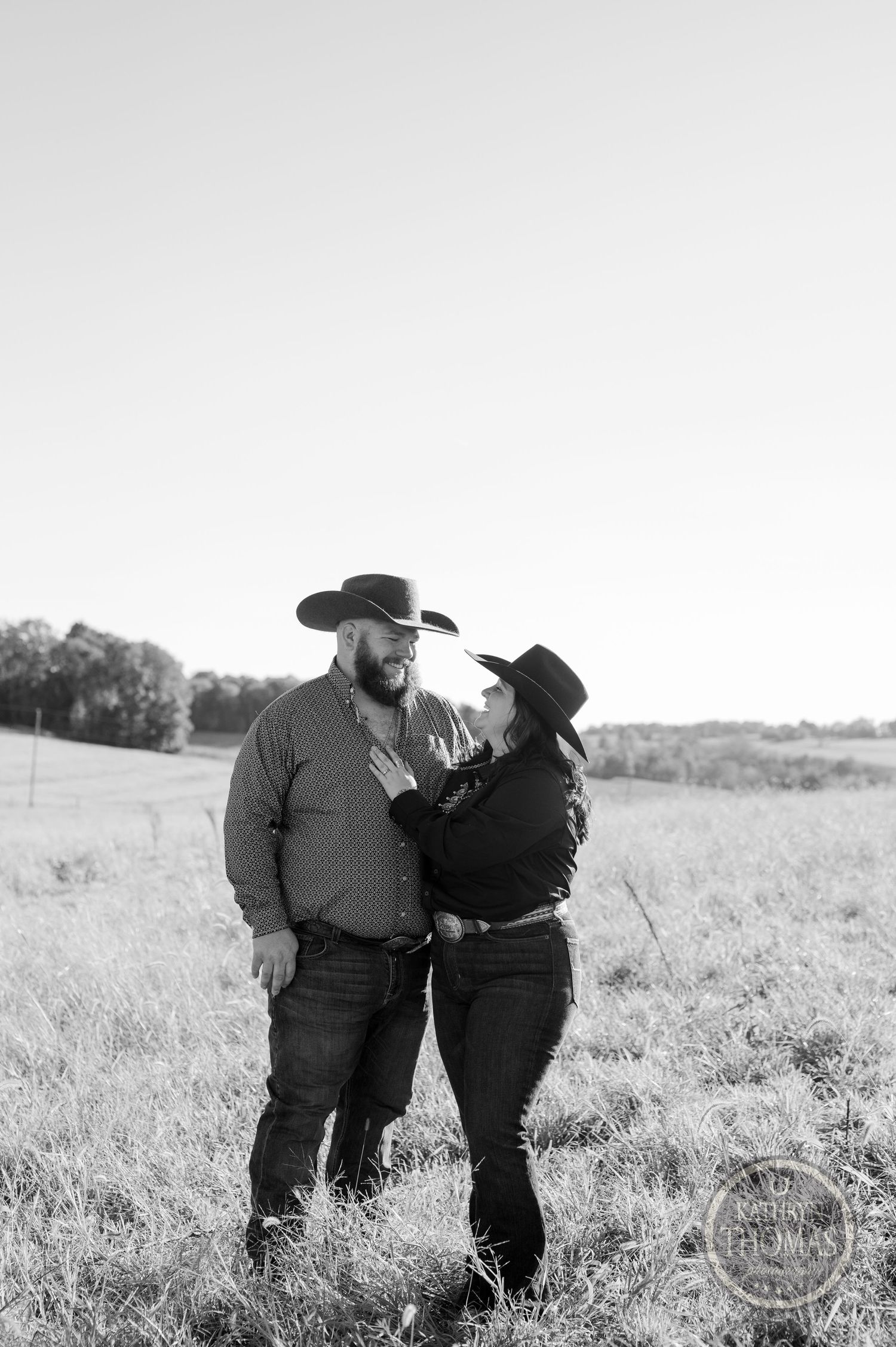 Black and white portrait of couple in western wear standing in grassy field.