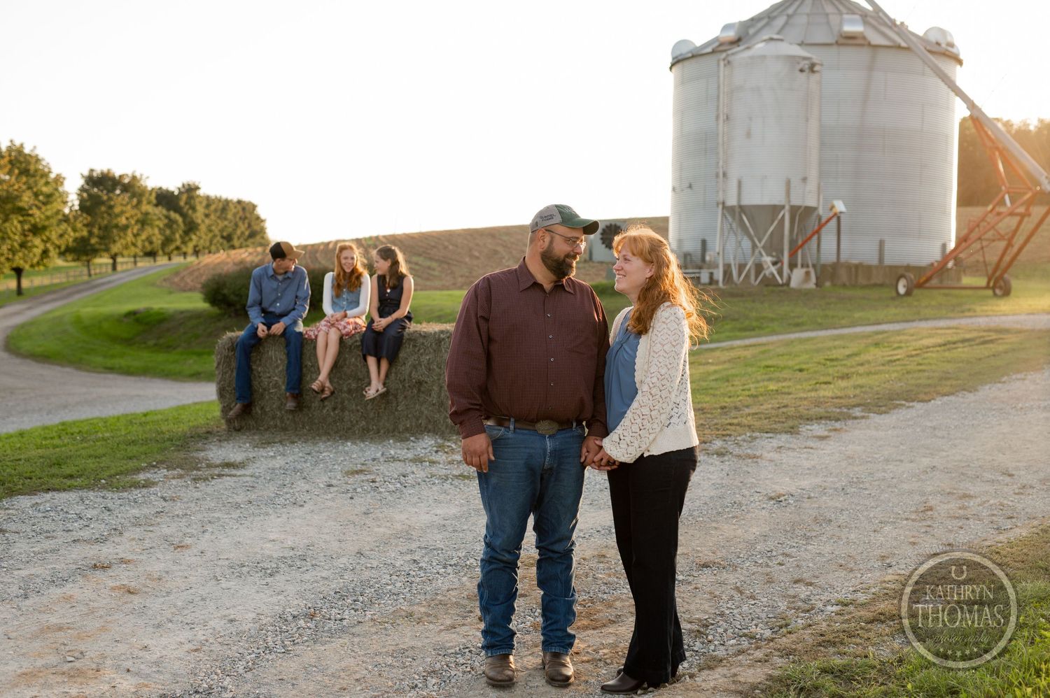 Couple poses in foreground with others sitting on hay bales in background by grain silo.