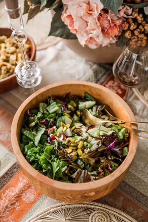 Fresh garden salad with mixed greens, nuts and seeds in wooden bowl alongside wine glasses and pink roses.