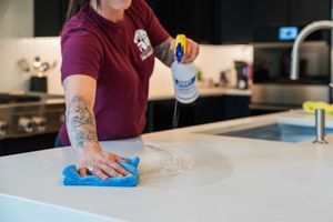Someone wearing a burgundy shirt wipes down a kitchen counter with a blue cleaning cloth.
