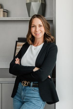 Professional woman in black blazer and jeans poses confidently against gray wall in modern office setting.