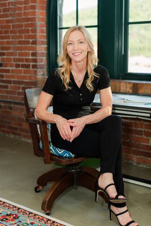 Professional in black attire sits confidently on office chair against exposed brick wall with natural lighting.
