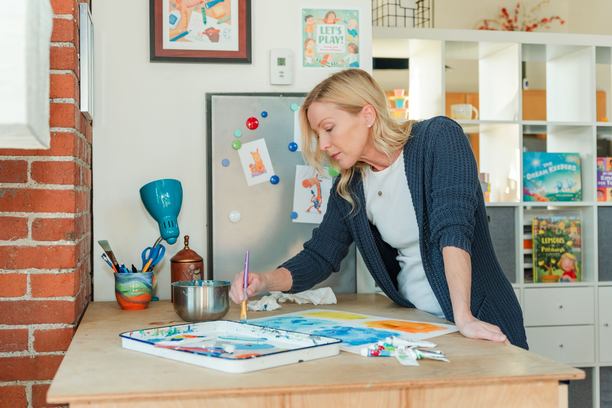 Creative artist leans over desk working on colorful artwork in bright home studio with artwork displayed on walls.