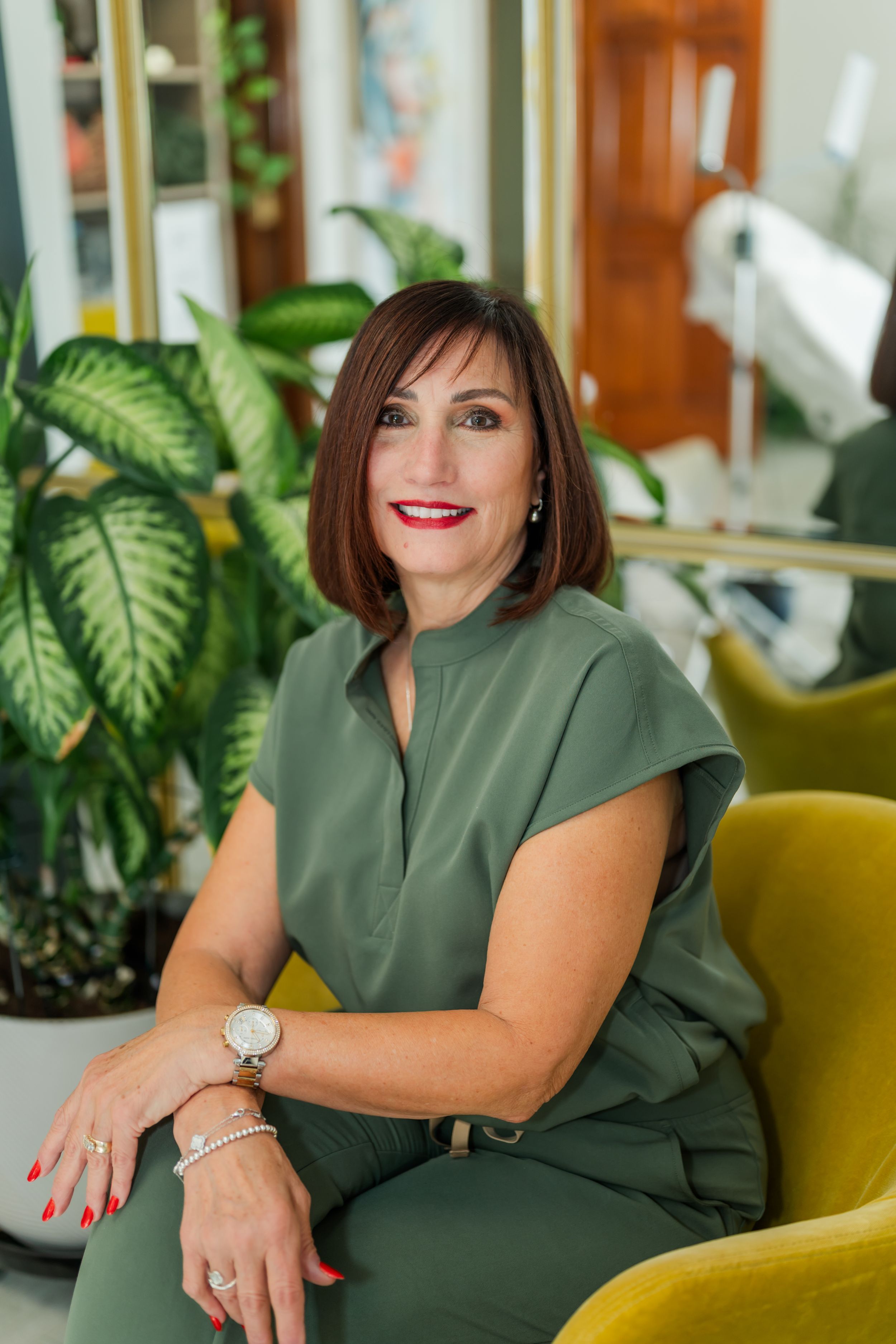 Professional portrait of someone in an olive green outfit seated in a yellow chair next to indoor plants.