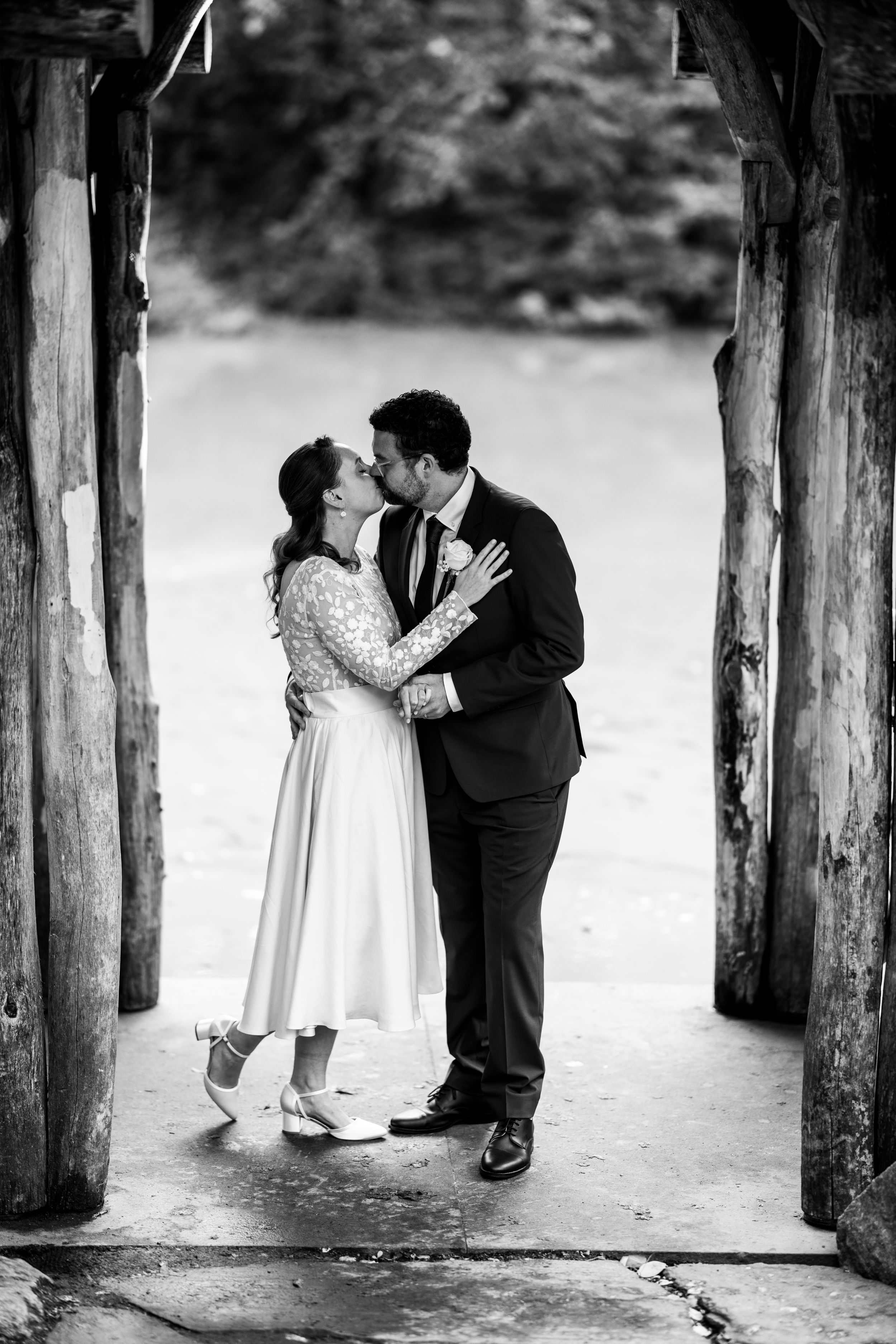 A romantic black and white wedding photo of a couple embracing under a rustic wooden archway.