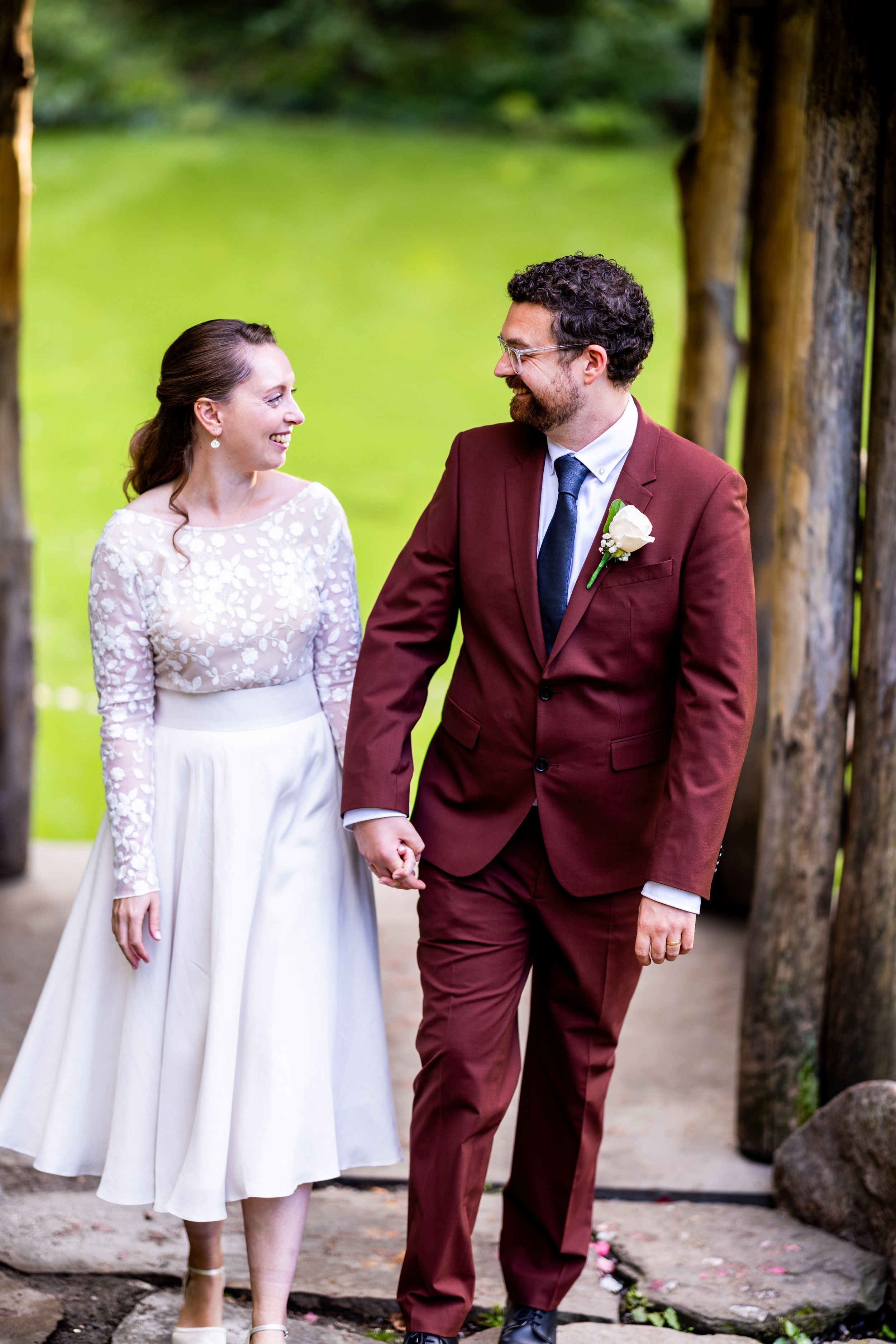 A couple in wedding attire sharing a joyful moment under a wooden pergola, with the groom wearing burgundy.