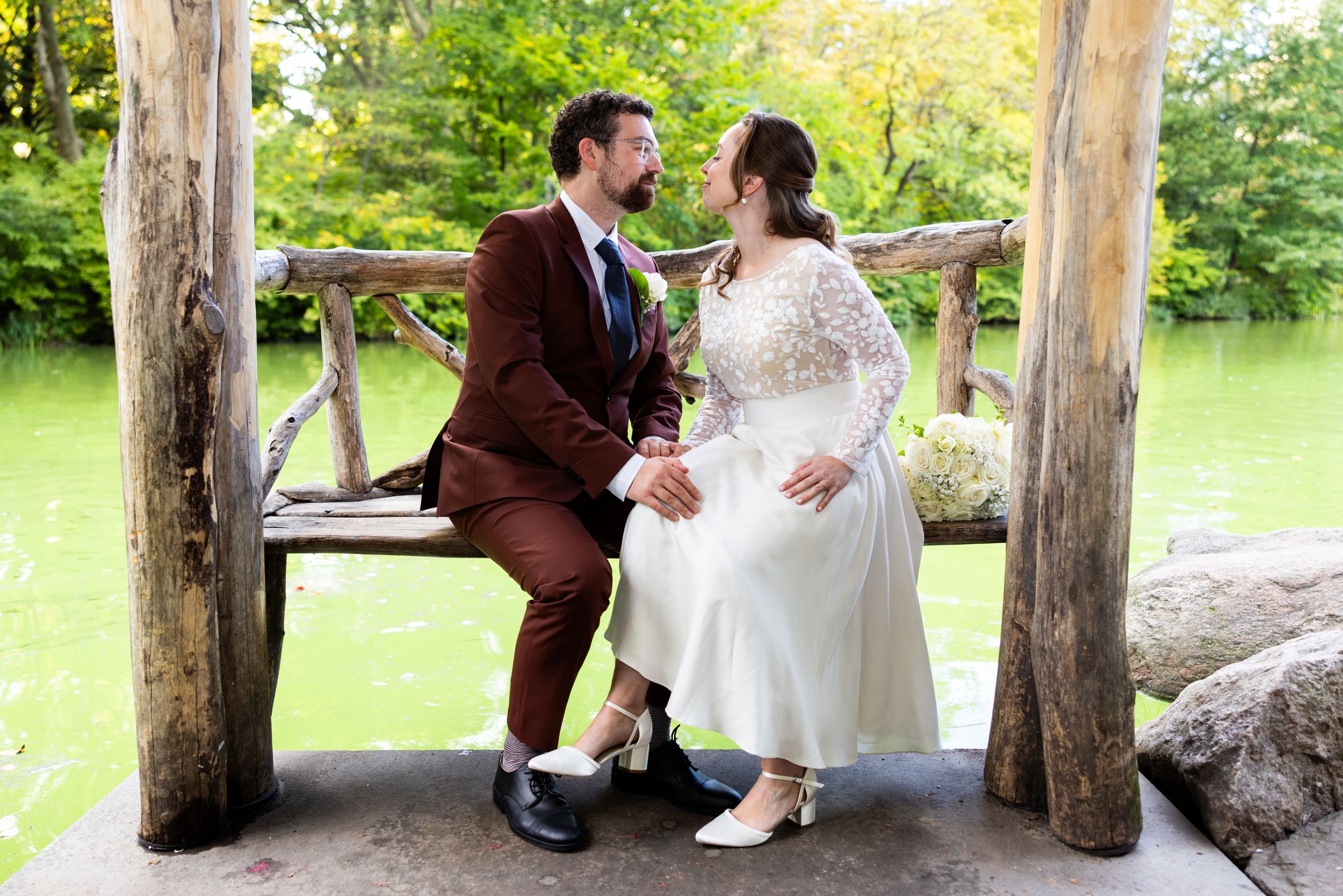 Newlyweds share intimate moment on rustic wooden bench in Wagners Cove Cental Park NYC during outdoor wedding.