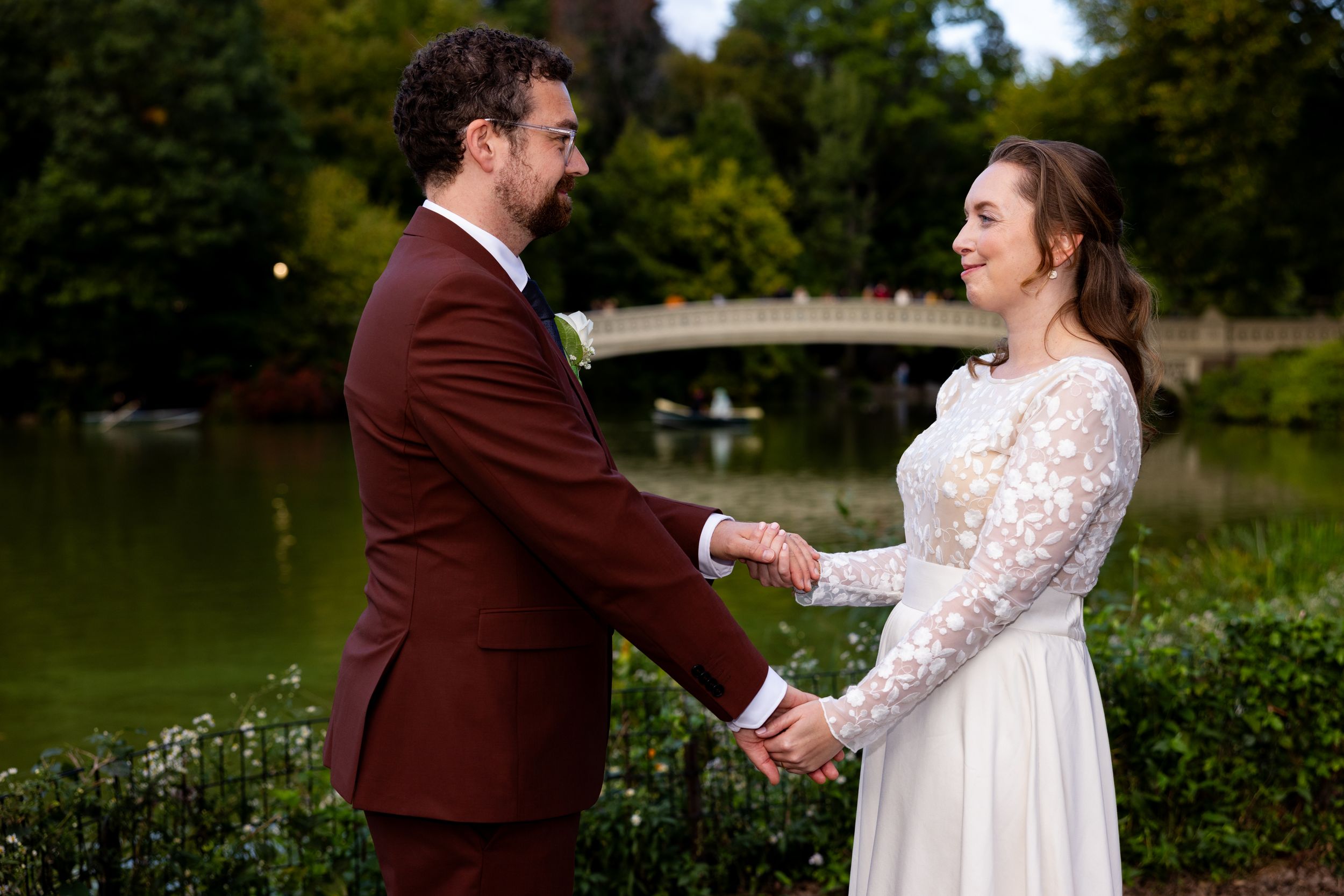 A couple holding hands in a garden setting in front of Bow Bridge Central Park NYC with the groom in burgundy and bride in lace.
