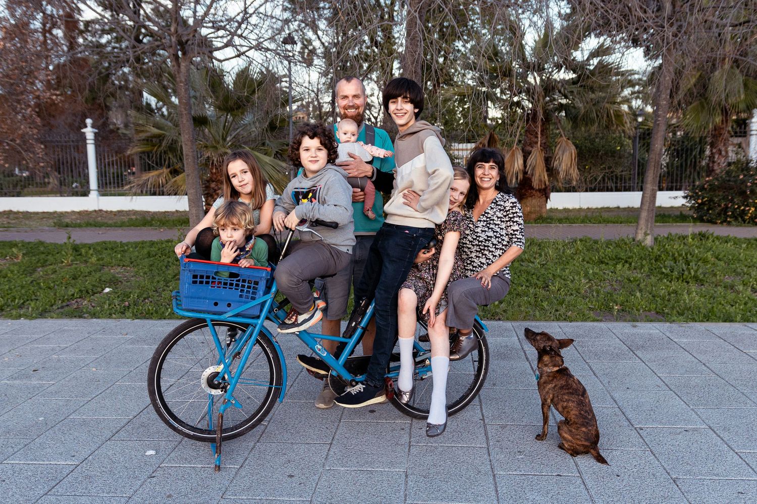 A group stands together with a cargo bike on a sidewalk while a brown dog sits nearby at dusk.