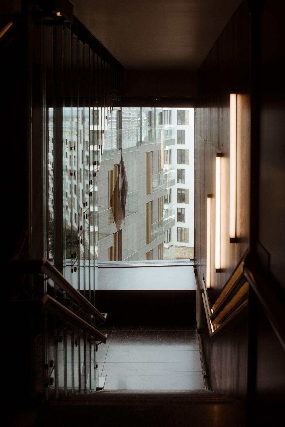 Dark stairwell interior with vertical fluorescent lights leading to a view of urban high-rise buildings.
