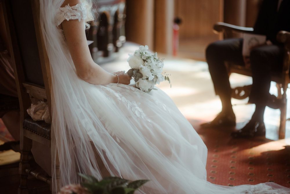 A bride in a flowing white dress holding a bouquet sits in soft window light.