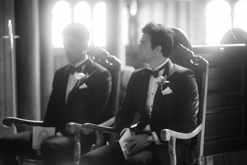 Two groomsmen in tuxedos sit in church pews during a wedding ceremony.