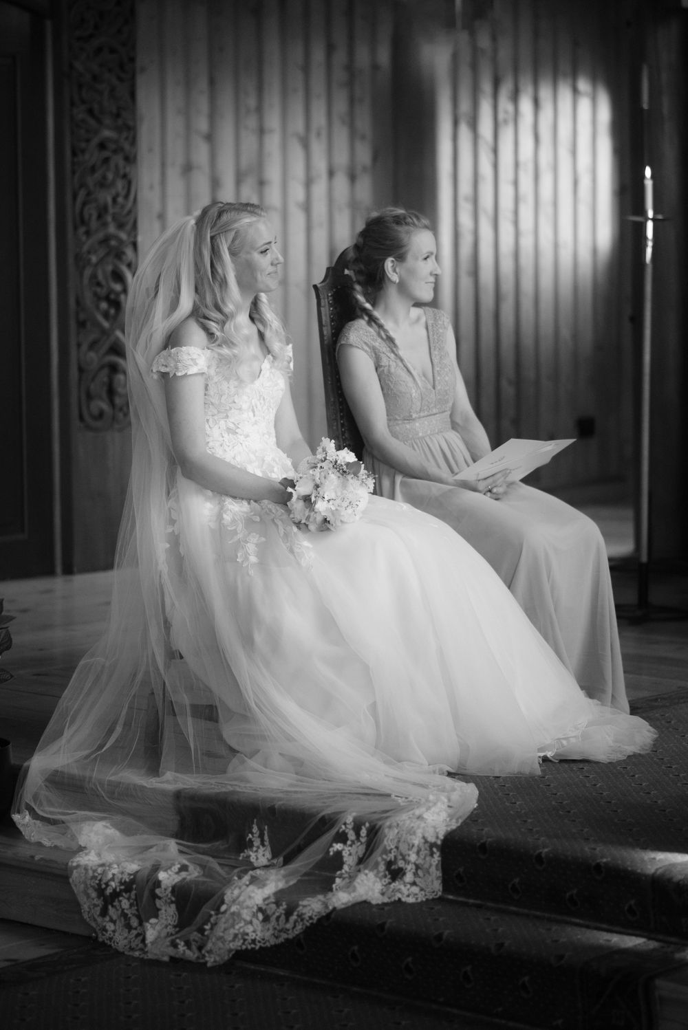 A bride and bridesmaid sit together in formal attire during a church ceremony.