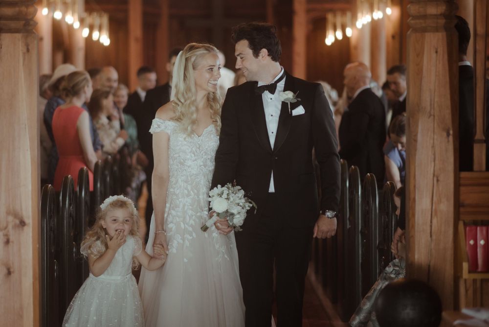 A wedding processional in a warmly lit church with guests looking on from wooden pews.