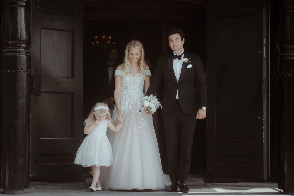 Elegant wedding portrait showing couple and young flowergirl in formal attire standing in church doorway.