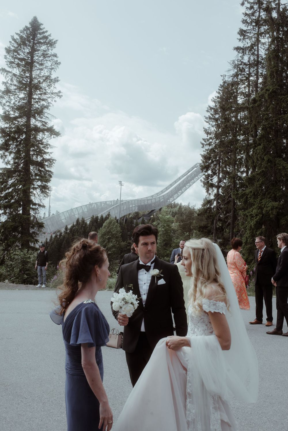Wedding party gathered outdoors with evergreen trees and ski jump in background.