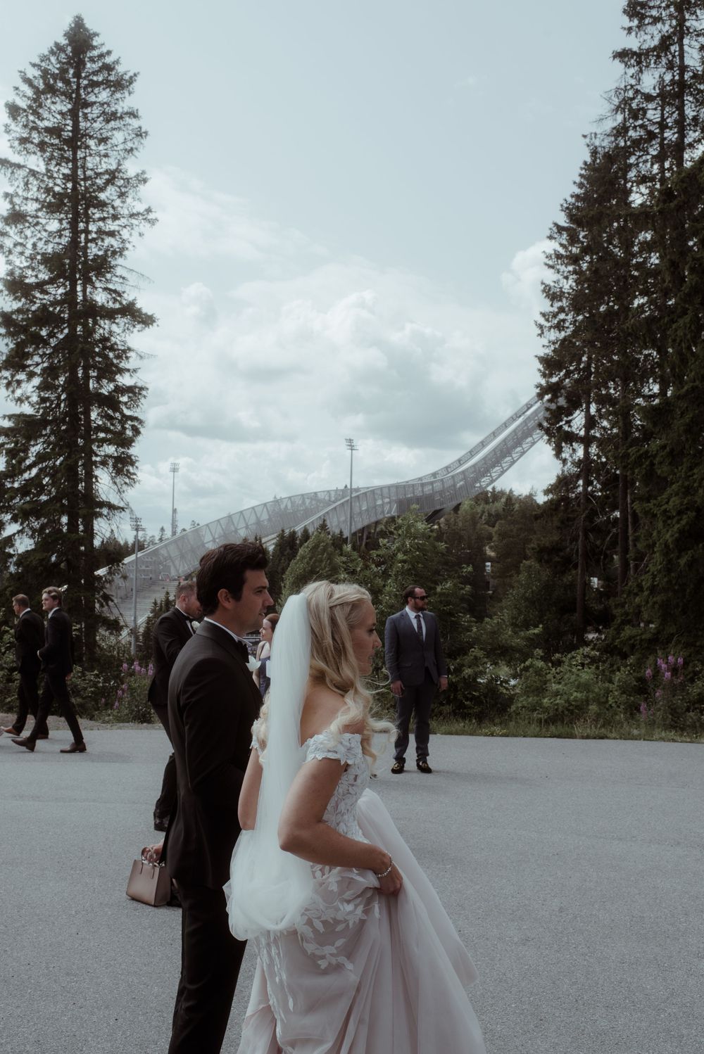 A wedding couple stands outdoors against a backdrop of tall evergreen trees and a ski jump slope.