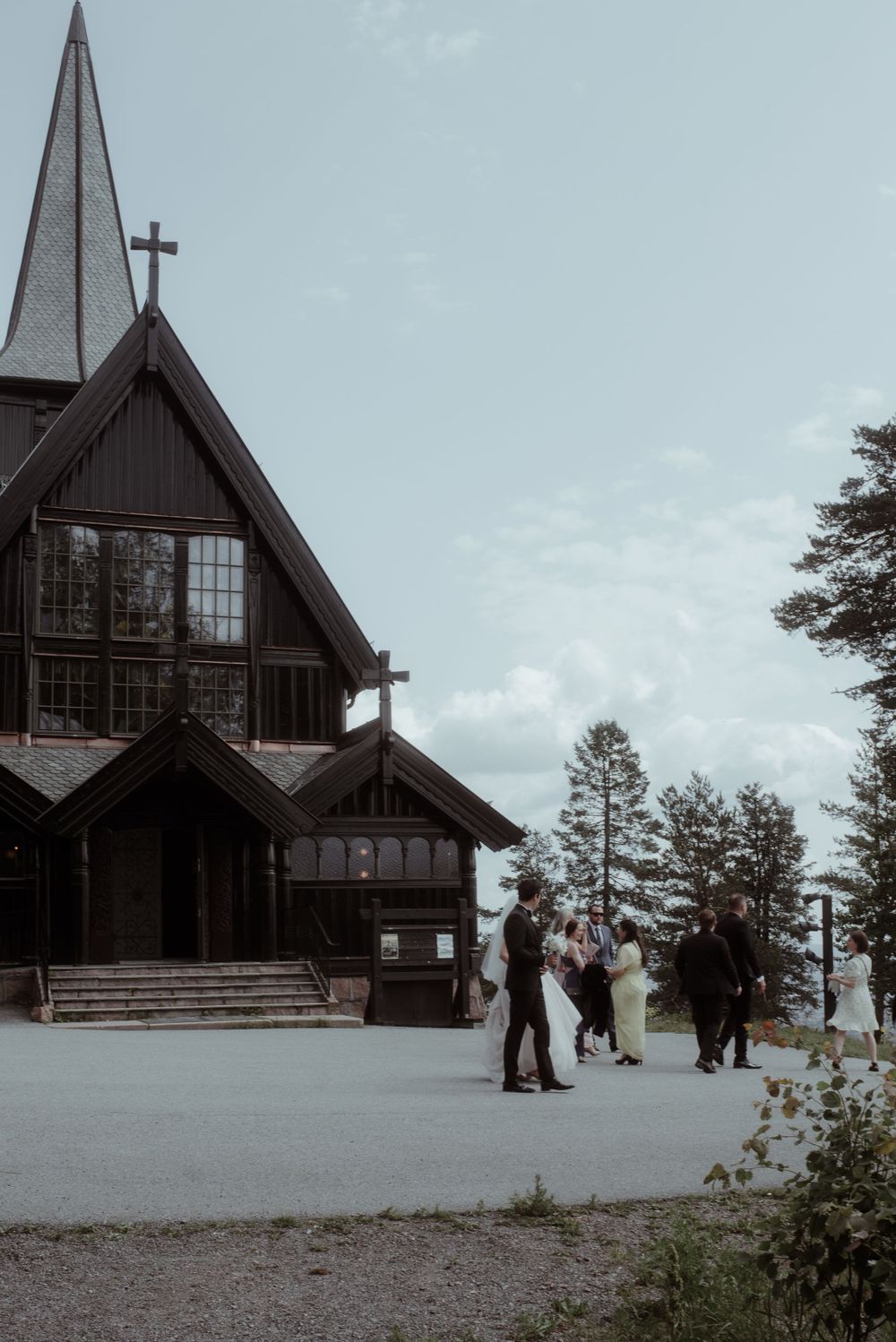 A wooden church with a tall spire stands against a cloudy sky as wedding guests gather outside.