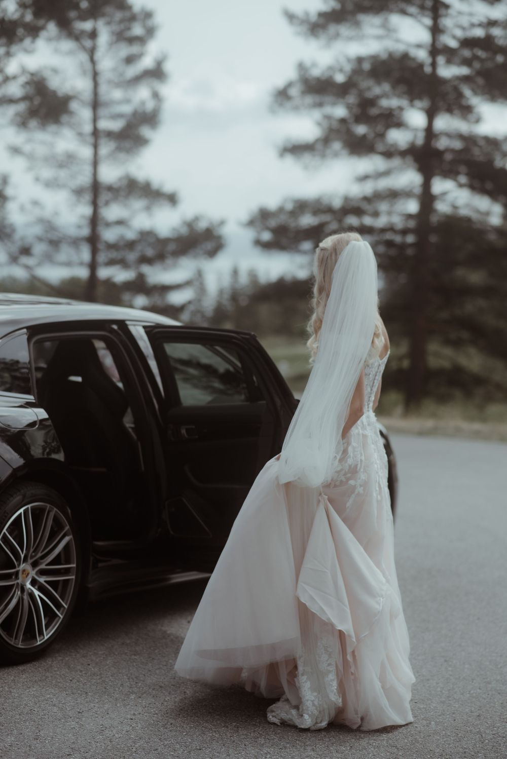 A bride in a flowing white dress stands beside a luxury car on a misty road.