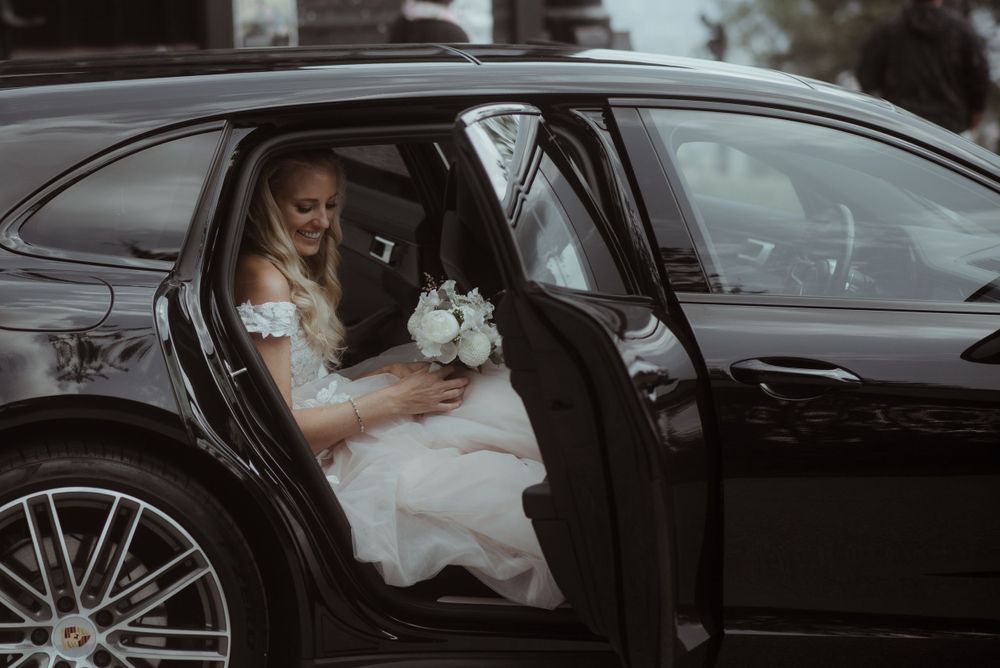A bride in a white dress sits in the backseat of a black luxury car holding a bouquet.