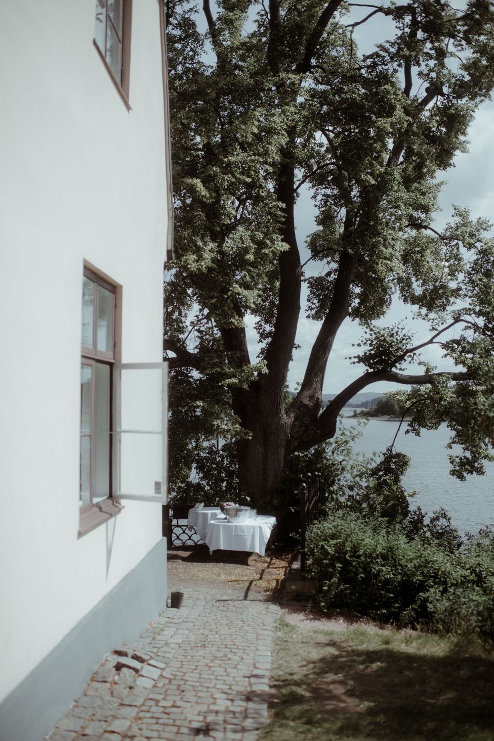 White building exterior with large tree and stone path leading to waterfront view.