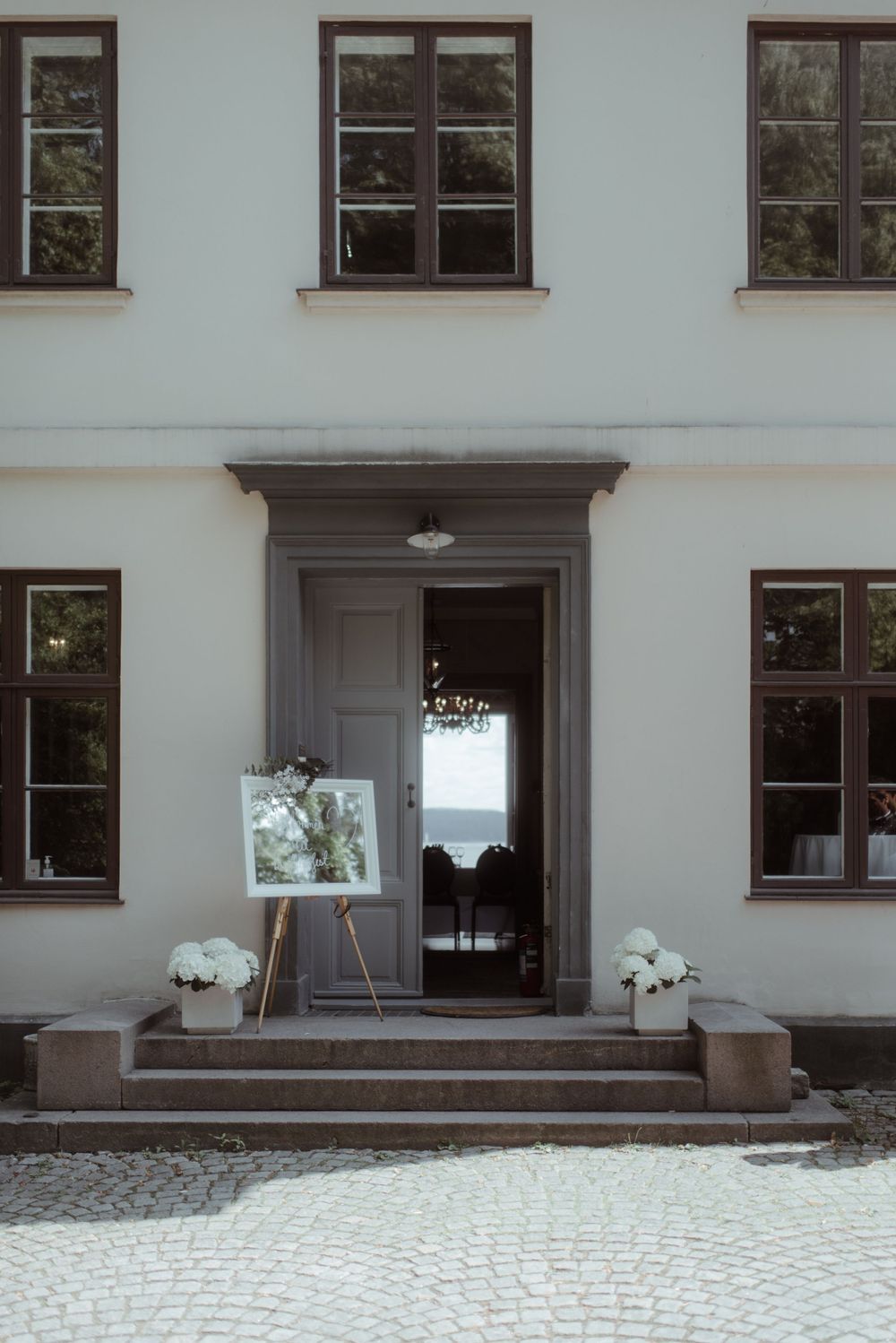 Classic white building entrance with grey door, windows, and potted flowers on steps.