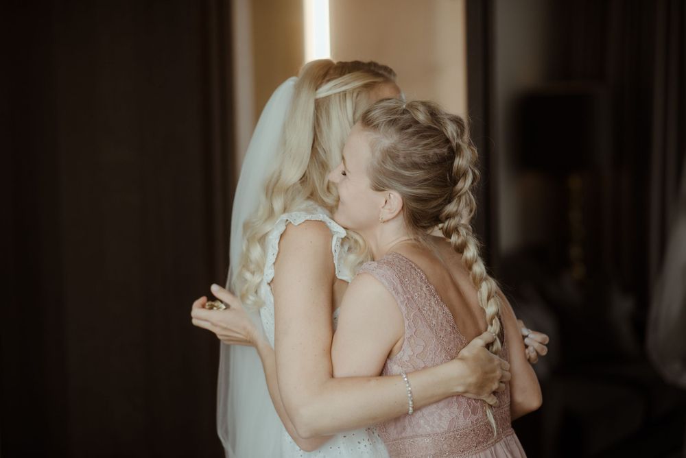 Two women share an emotional embrace during wedding preparations in a softly lit room.