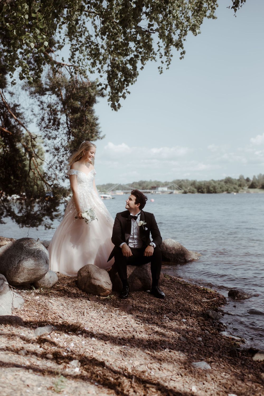 A wedding couple shares a tender moment by the waterfront under hanging tree branches.