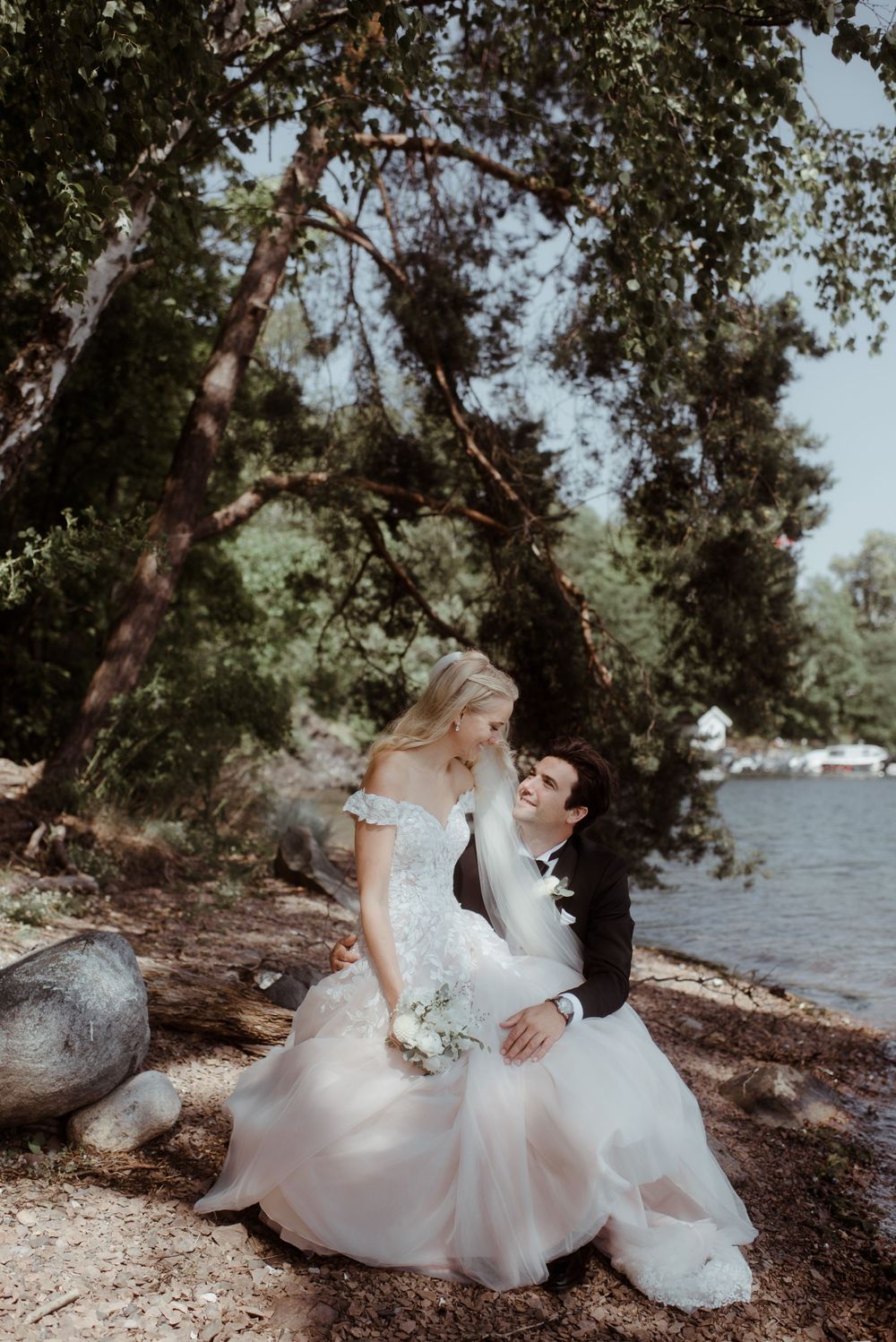Newlyweds in formal attire sit together on a rocky shoreline under overhanging trees.