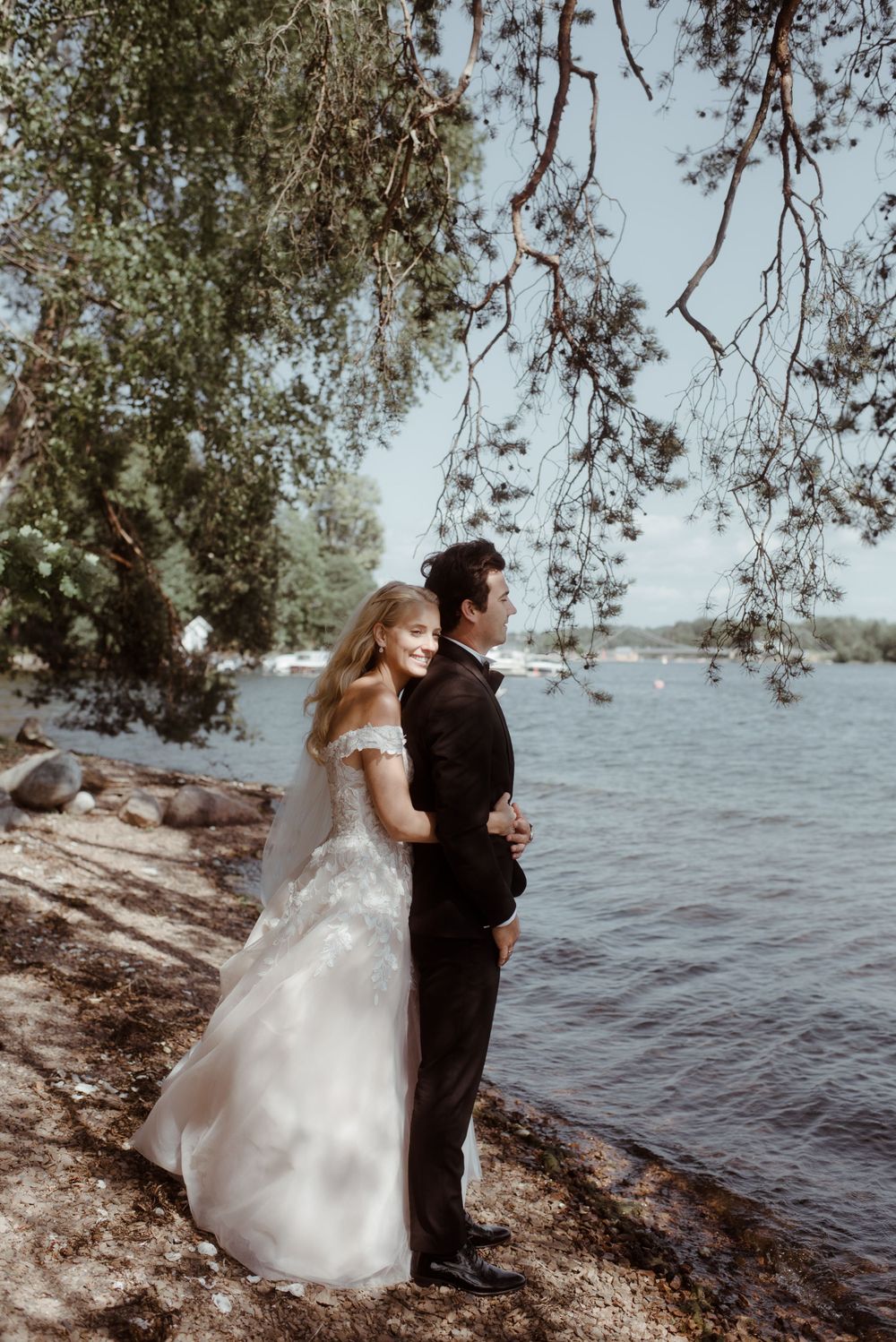A bride and groom pose together on the shoreline under hanging tree branches.