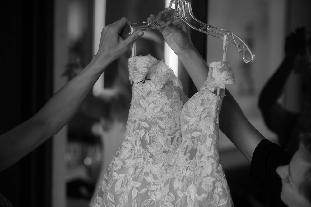 Multiple hands hold up an ornate white lace wedding dress in black and white.