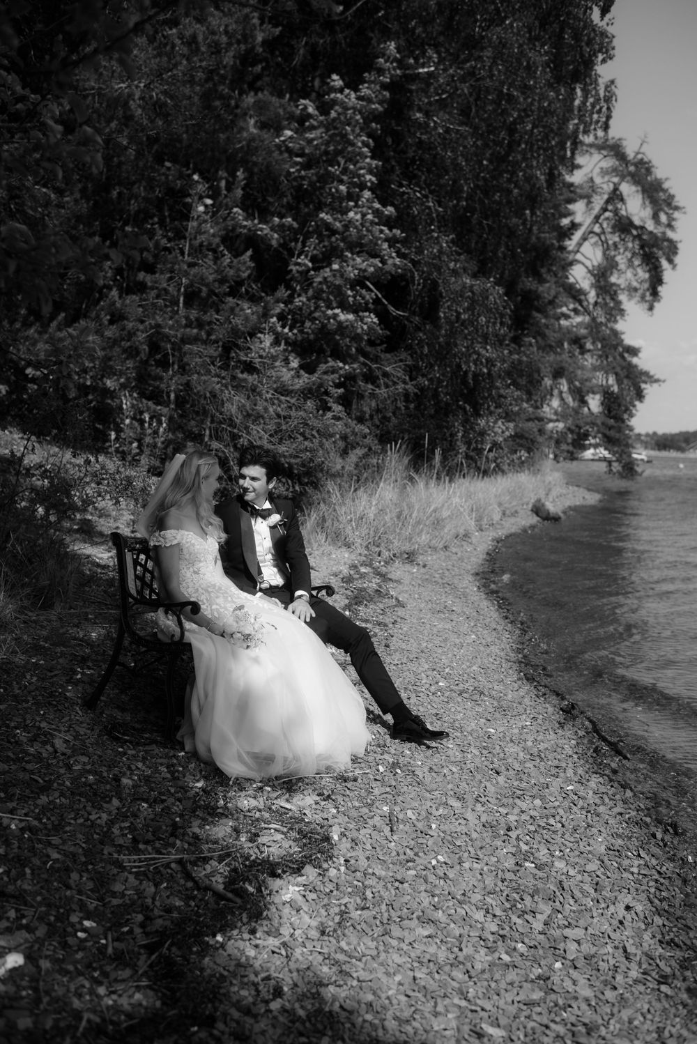 A black and white photo of newlyweds sitting on a bench along a lakeside path.