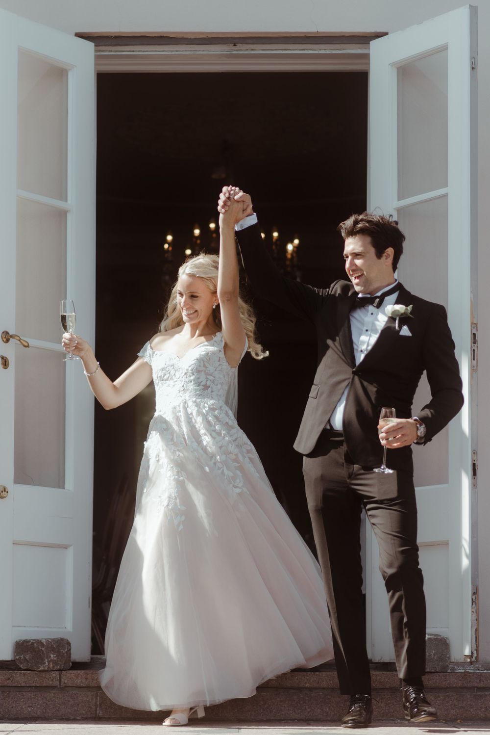 A newlywed couple celebrates joyfully in their wedding attire while exiting through white doors.