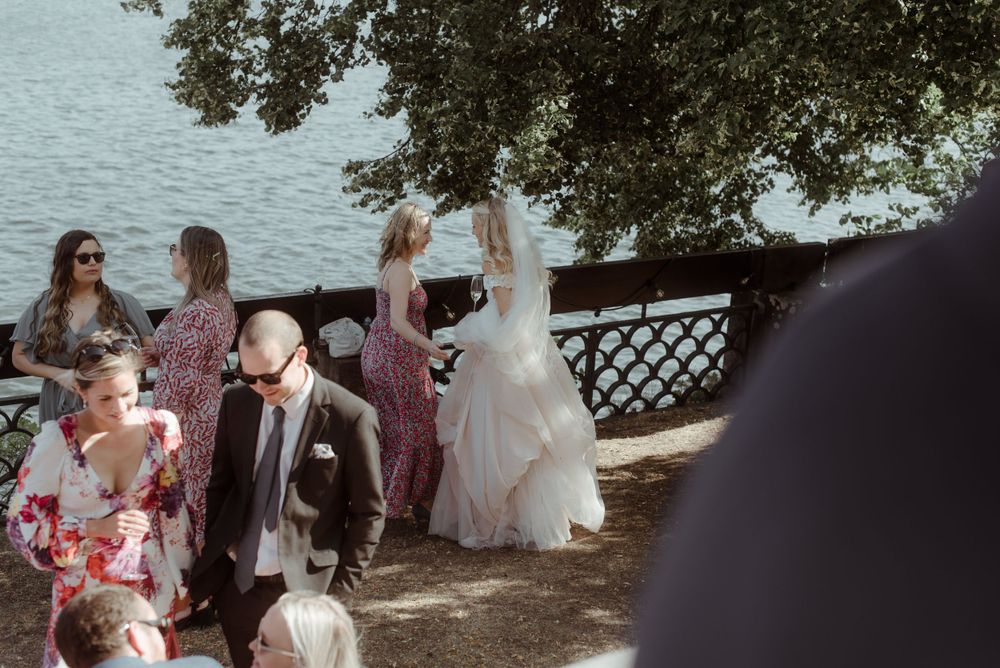 Wedding guests gather outdoors near a waterfront railing under shady trees.