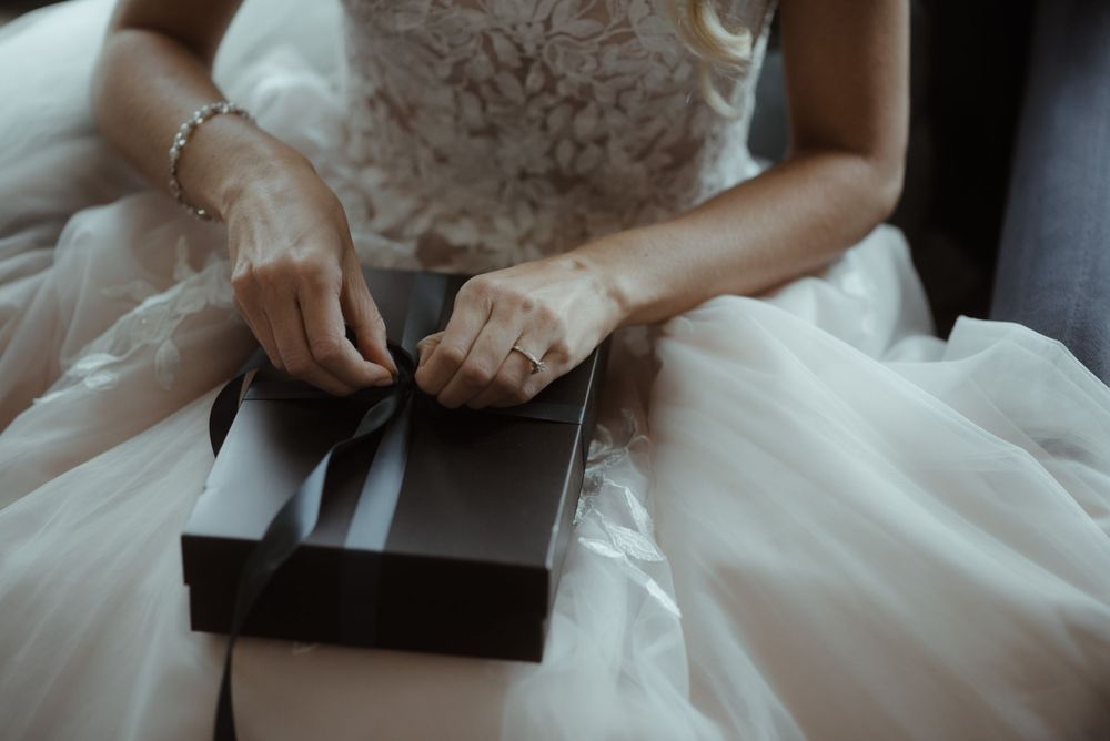 Bride in white dress opening a small dark box while seated.