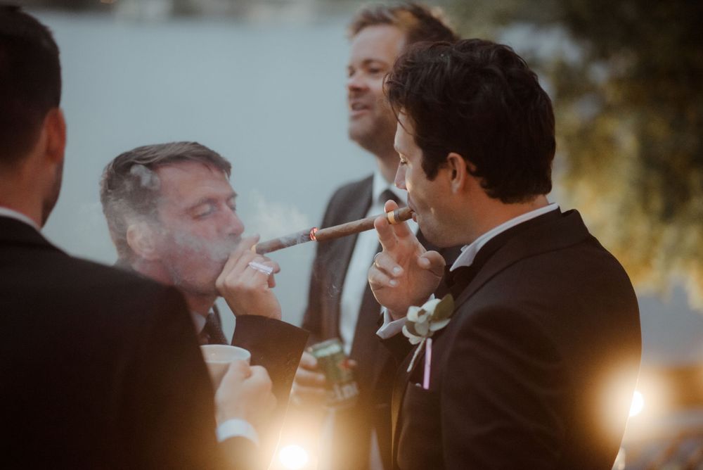 Wedding guests smoking cigars and chatting during sunset at an outdoor reception.