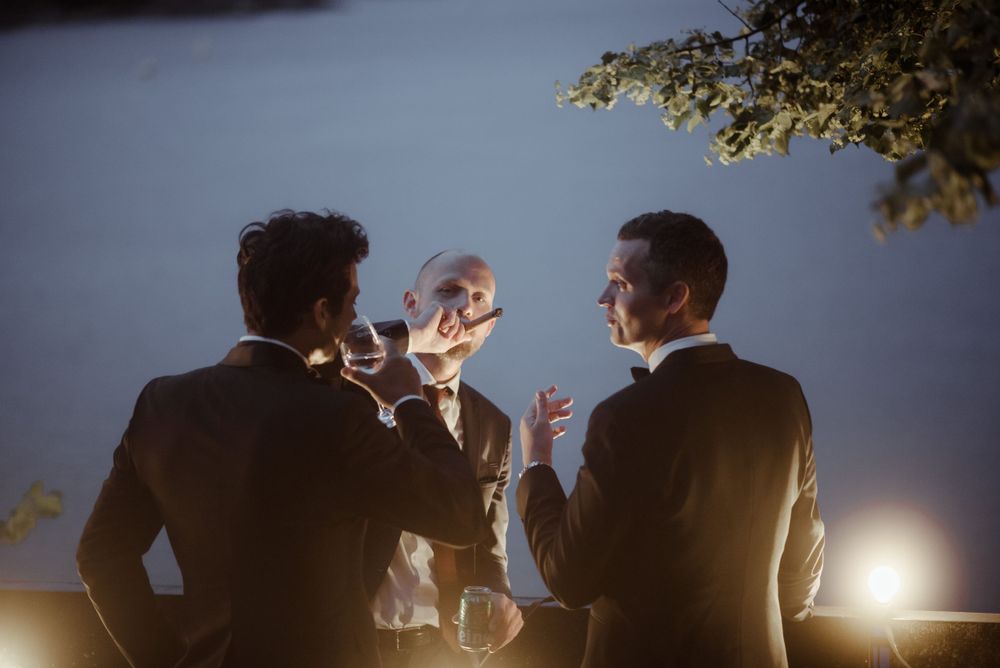 Silhouettes of guests enjoying drinks at dusk during an outdoor wedding celebration.