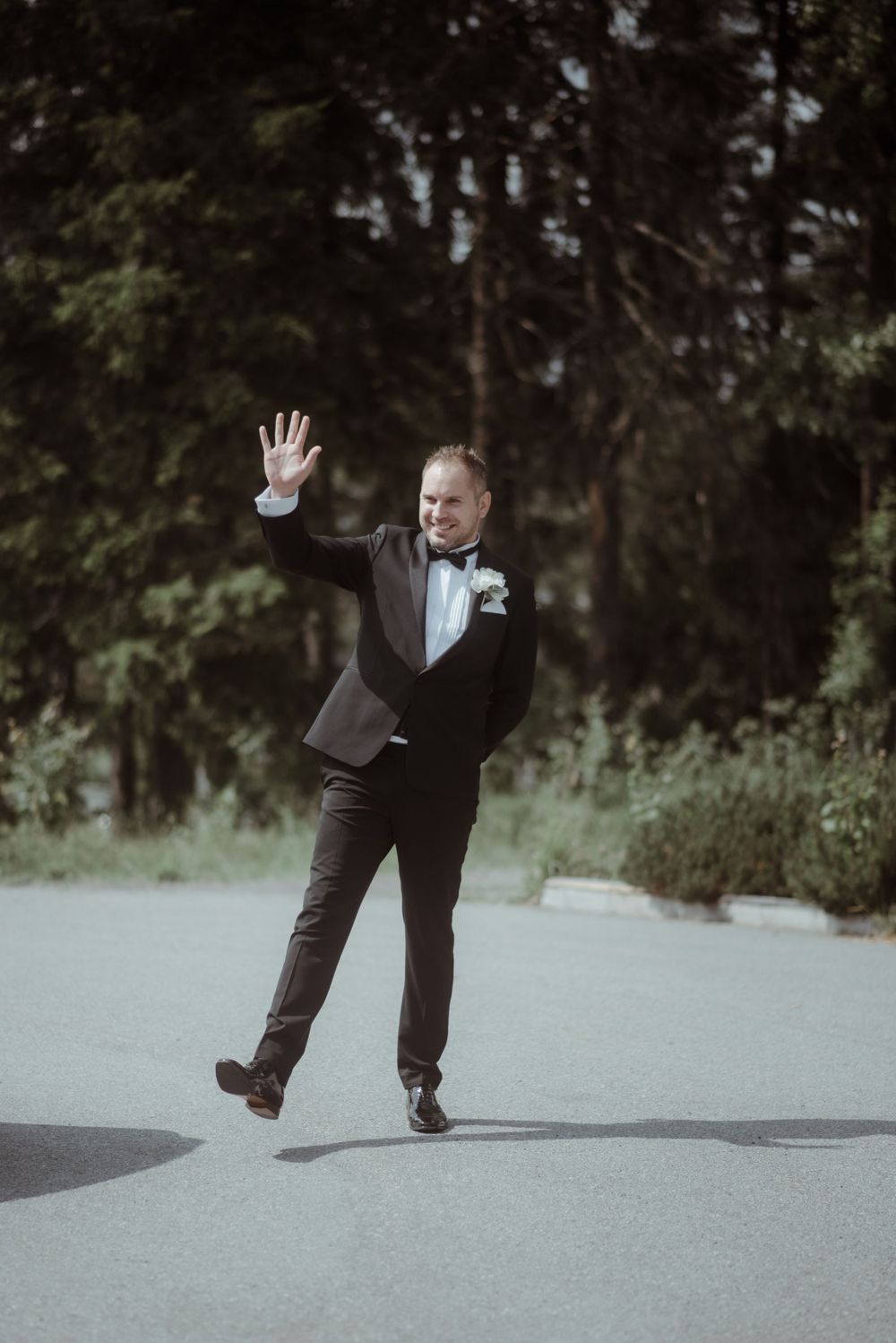 A groom in a classic tuxedo strikes a playful pose outdoors.
