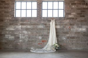 Elegant white wedding dress hangs against rustic brick wall beneath industrial windows with bouquet accent.